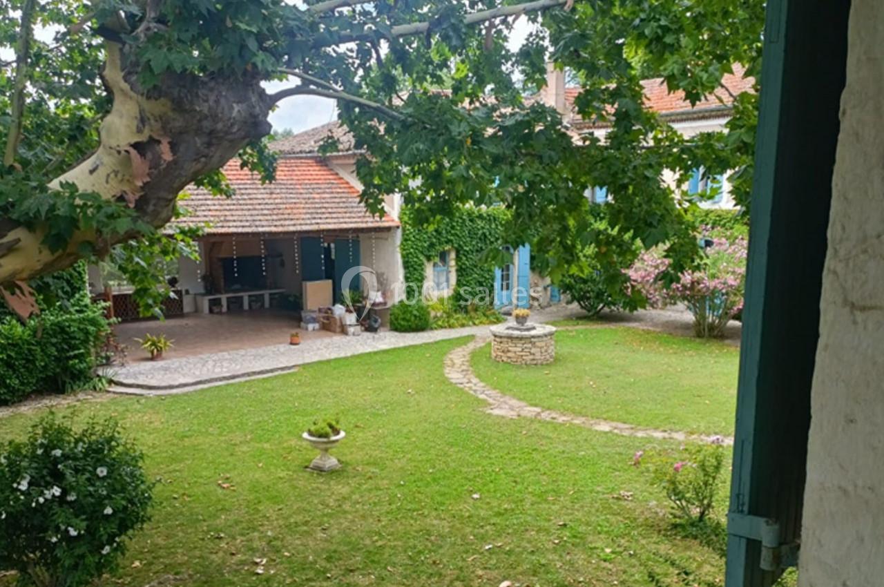 Cour intérieure verdoyante avec pelouse, arbre, fontaine en pierre et maison en arrière-plan sous un ciel partiellement…