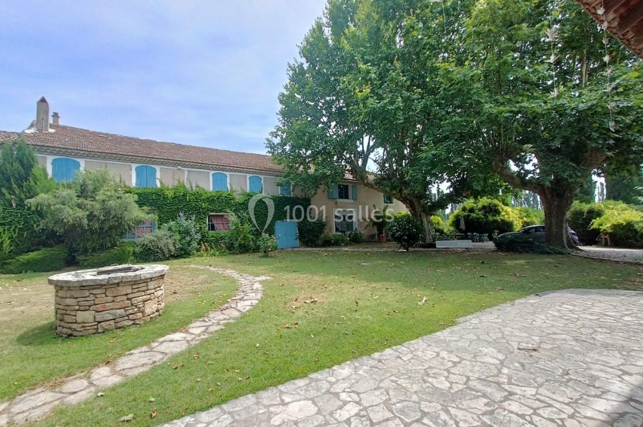 Grande maison en pierre avec volets bleus, entourée d'un jardin arboré, d'un puits en pierre et d'une allée pavée.