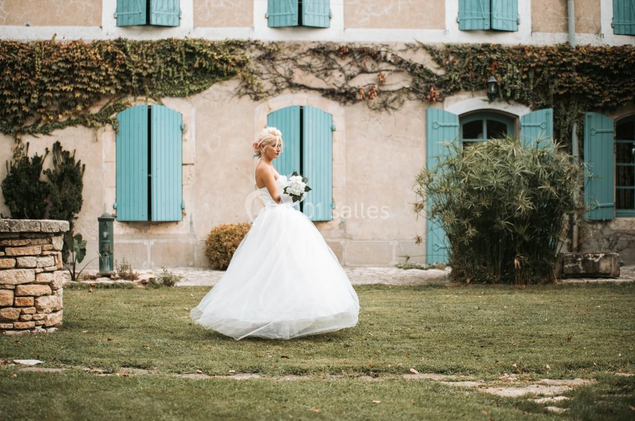 Une mariée en robe blanche tient un bouquet dans un jardin devant une maison aux volets bleus.