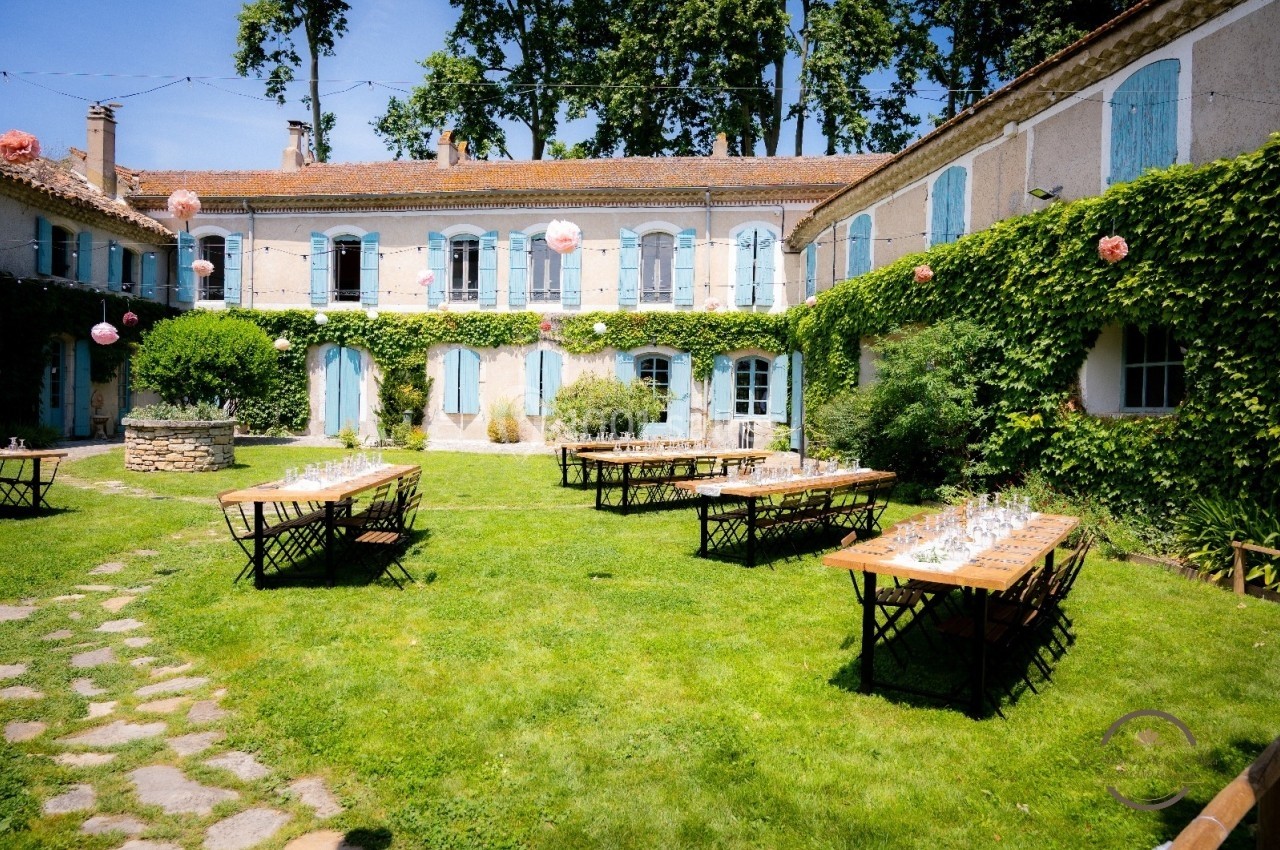 Cour intérieure verdoyante avec tables en bois dressées, entourée de bâtiments anciens aux volets bleus.