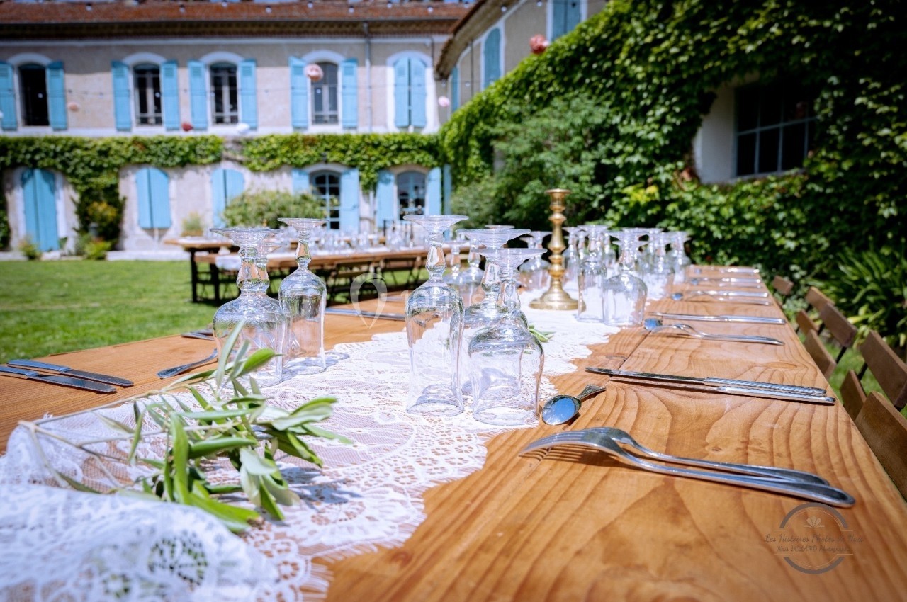 Grande table en bois dressée avec nappes en dentelle, verres et couverts, dans un jardin devant une maison aux volets bleus.