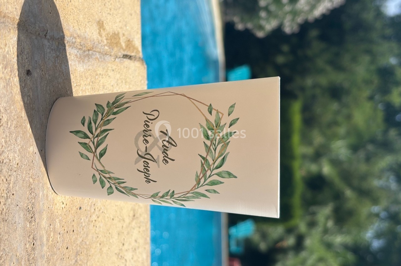 Livret beige décoré d'une couronne végétale, posé sur une surface en pierre près d'une piscine entourée de verdure.