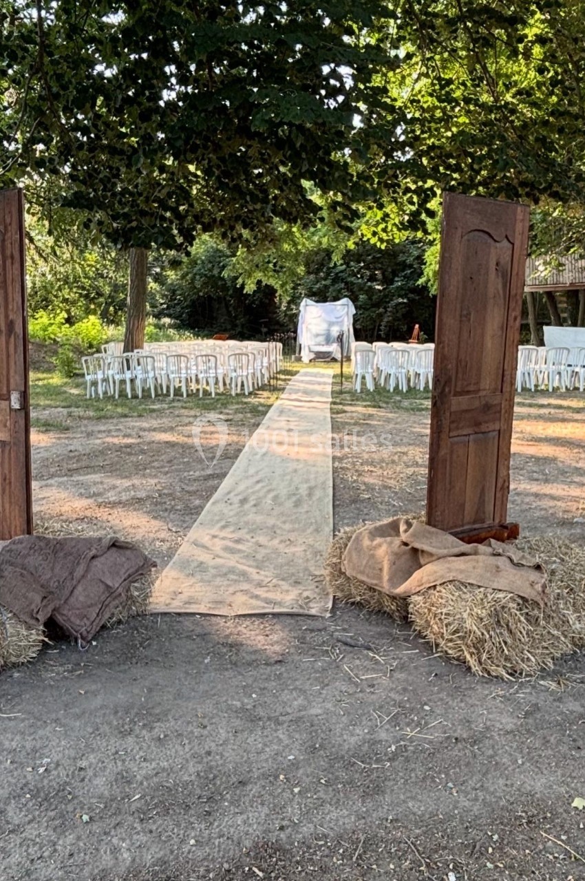 Allée centrale en extérieur bordée de chaises blanches, encadrée par deux portes en bois et des bottes de foin.
