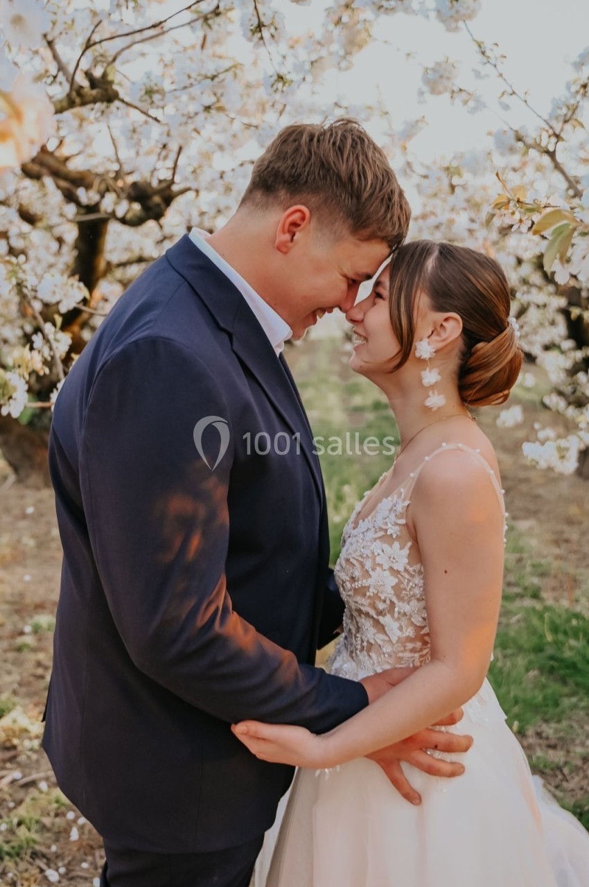 Un couple en tenue de mariage se tient face à face sous des arbres en fleurs, échangeant un moment tendre.
