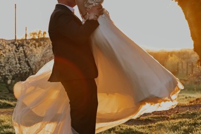 Un homme en costume soulève une femme en robe blanche dans un jardin au coucher du soleil.