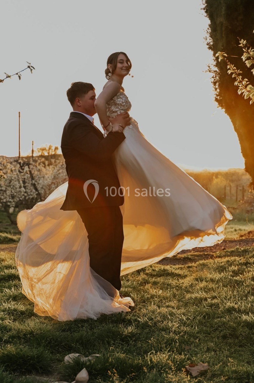 Un homme en costume soulève une femme en robe blanche dans un jardin au coucher du soleil.