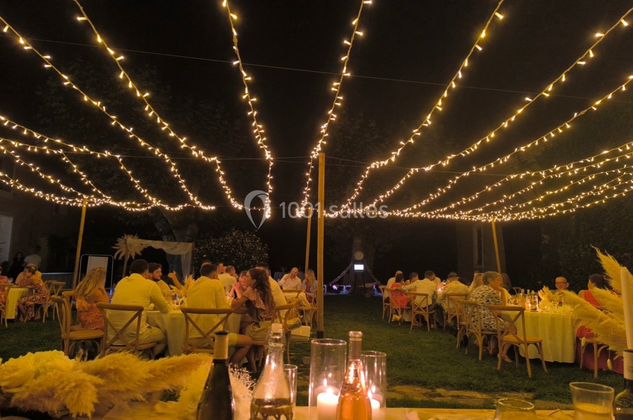 Dîner en plein air sous des guirlandes lumineuses, avec des invités assis à des tables rondes dans un jardin de nuit.