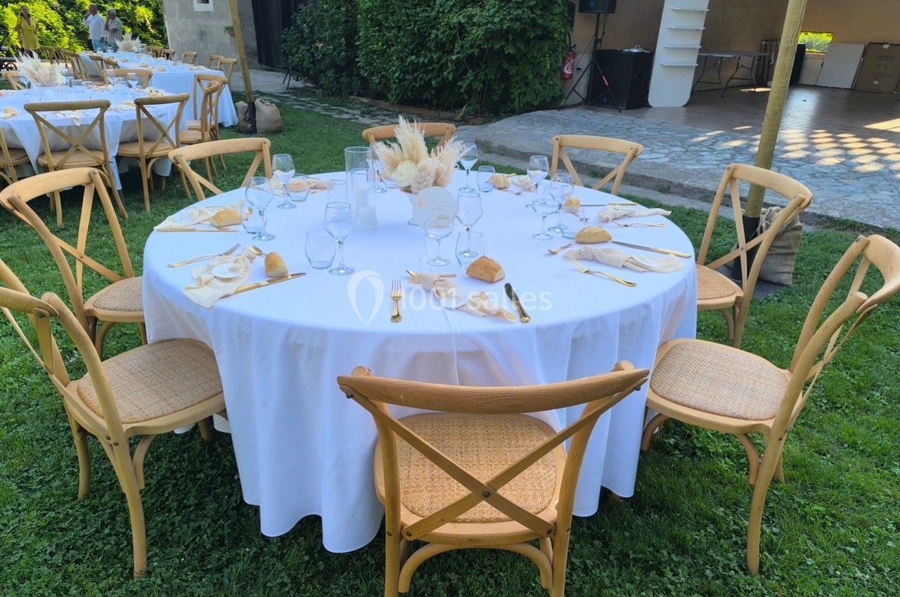 Table ronde dressée pour un repas en extérieur, avec nappes blanches, couverts dorés et décoration florale centrale.