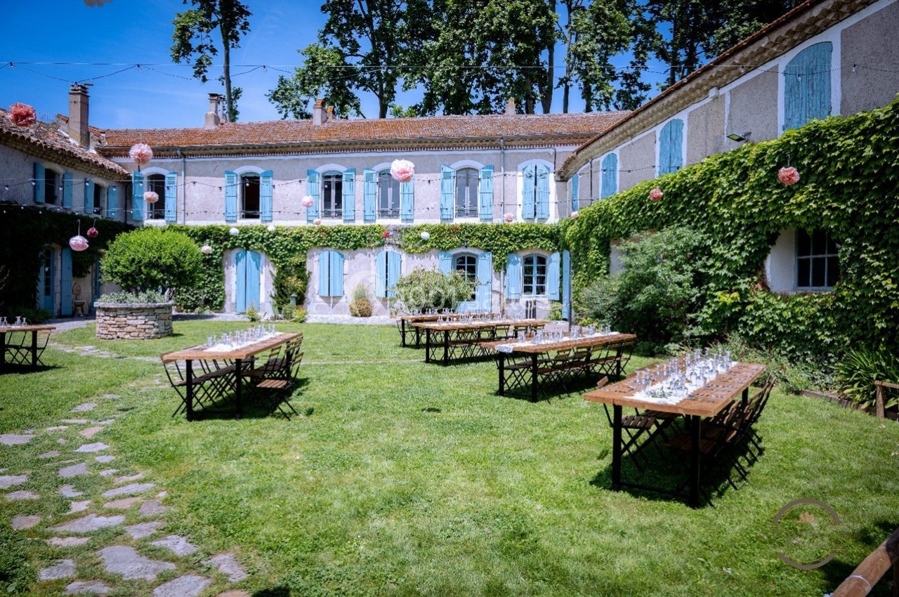 Cour verdoyante avec tables en bois dressées pour un événement, entourée d'un bâtiment ancien aux volets bleus.