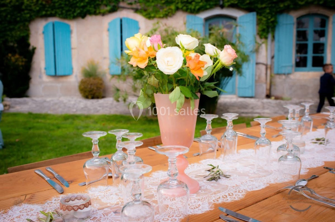 Vase de fleurs colorées sur une table en bois décorée, devant une maison en pierre aux volets bleus.