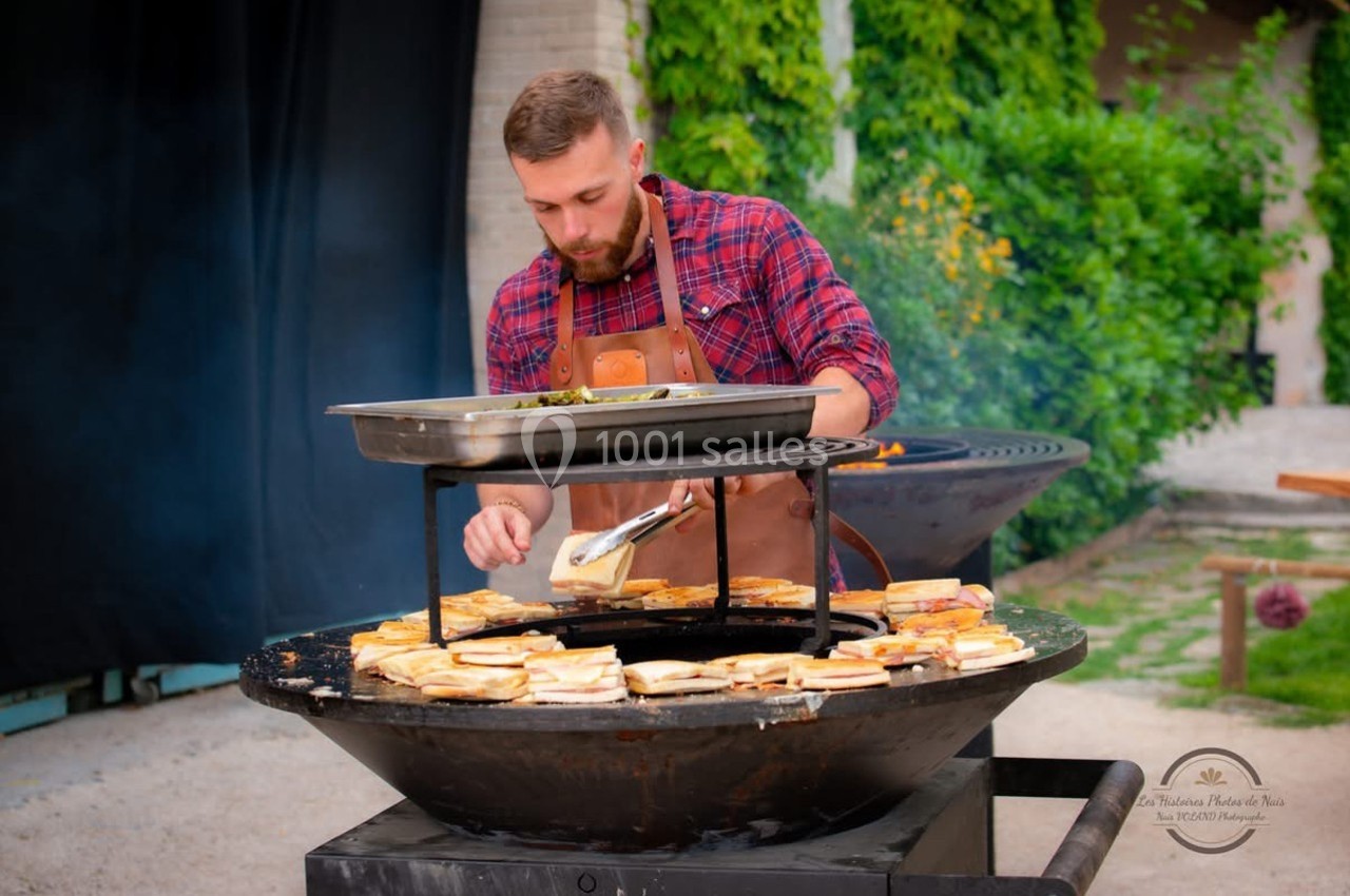 Un homme en tablier cuisine des sandwichs sur un grand barbecue rond en extérieur, entouré de verdure.