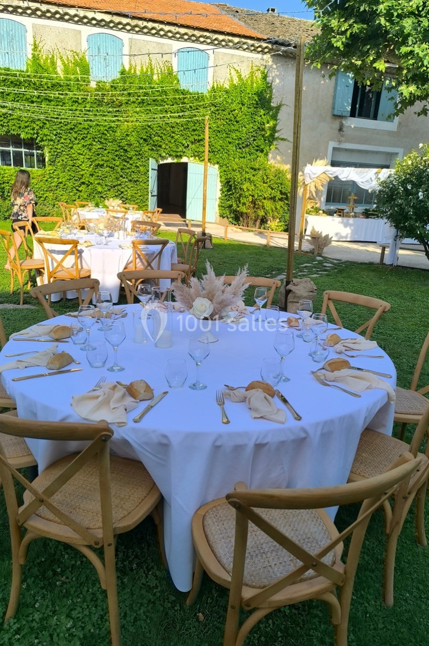 Table ronde dressée pour un repas en extérieur, avec nappes blanches, chaises en bois et décorations florales.