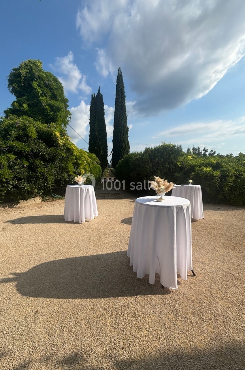 Trois tables hautes drapées de nappes blanches disposées en extérieur, entourées de verdure et de cyprès.