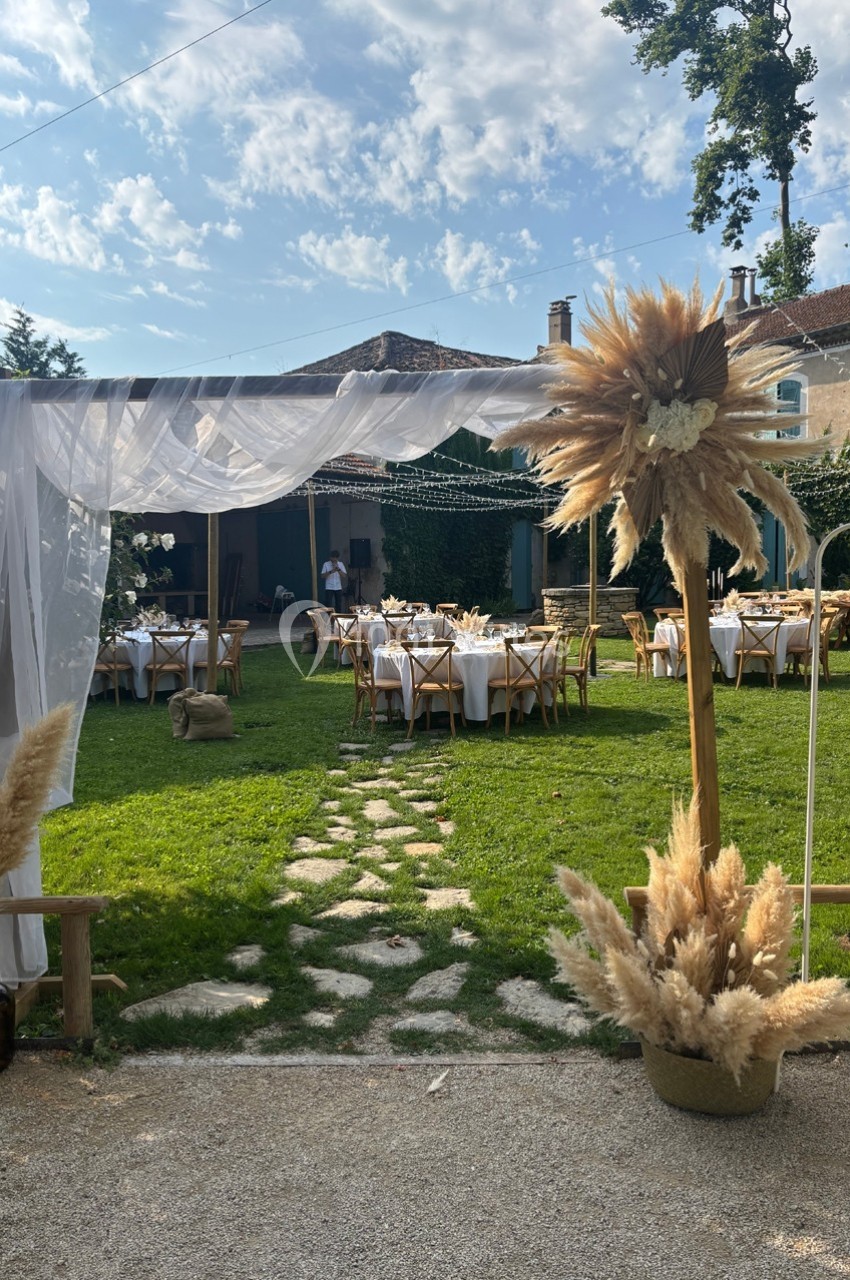 Décoration de mariage en extérieur avec arche en voilage blanc, tables dressées et ornements de pampas sur pelouse.