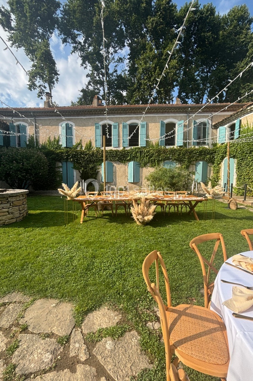 Grande table en bois décorée de fleurs séchées dans un jardin, devant une maison en pierre aux volets bleus.