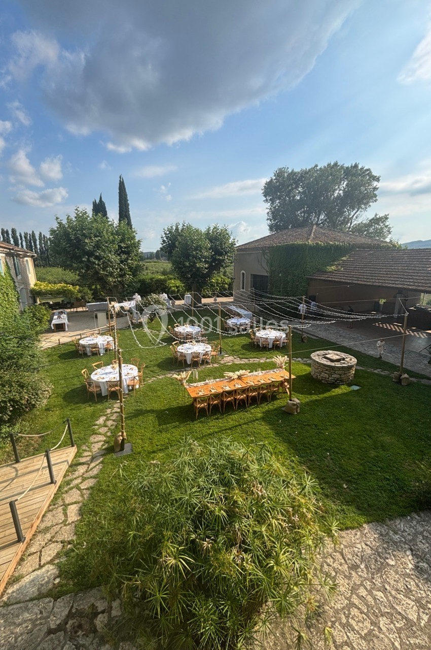 Jardin aménagé pour un événement avec tables dressées, chaises en bois, guirlandes lumineuses et puits central.