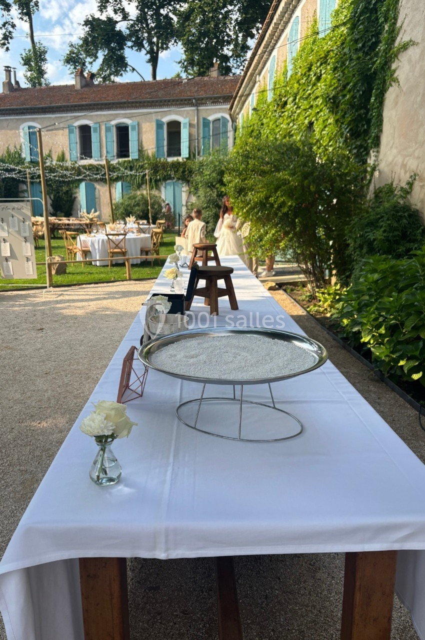 Grande table extérieure dressée avec une nappe blanche, décorée de fleurs, dans une cour ensoleillée près d'un bâtiment…