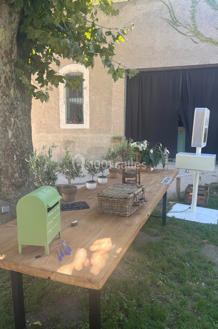 Table en bois à l'extérieur avec des plantes en pot, une boîte aux lettres verte, des paniers et un photomaton en arrière…