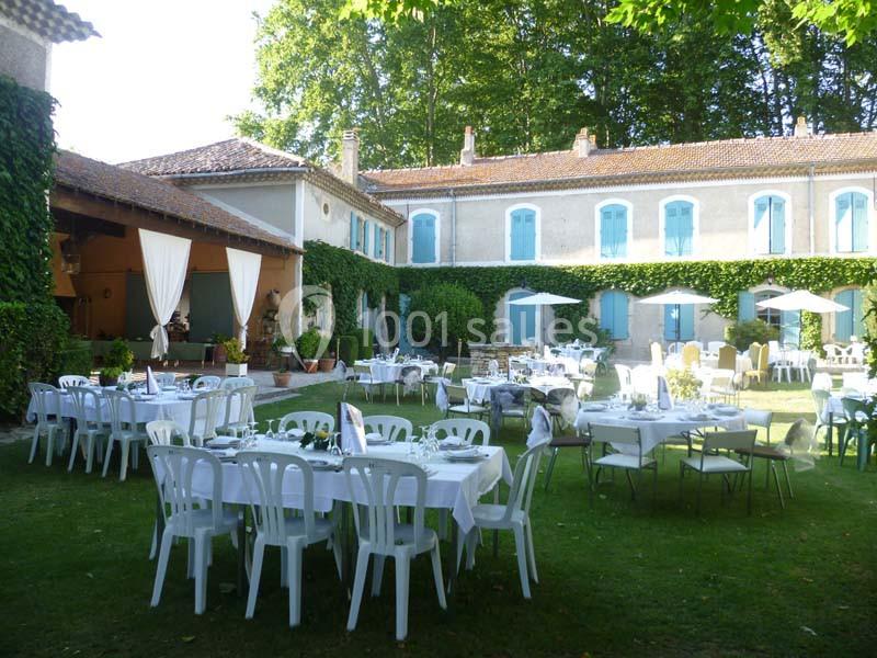 Tables et chaises blanches disposées dans un jardin verdoyant devant un bâtiment ancien avec volets bleus.