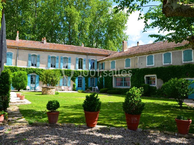 Grande maison de campagne avec façade en pierre, volets bleus, jardin verdoyant et pots de fleurs en premier plan.