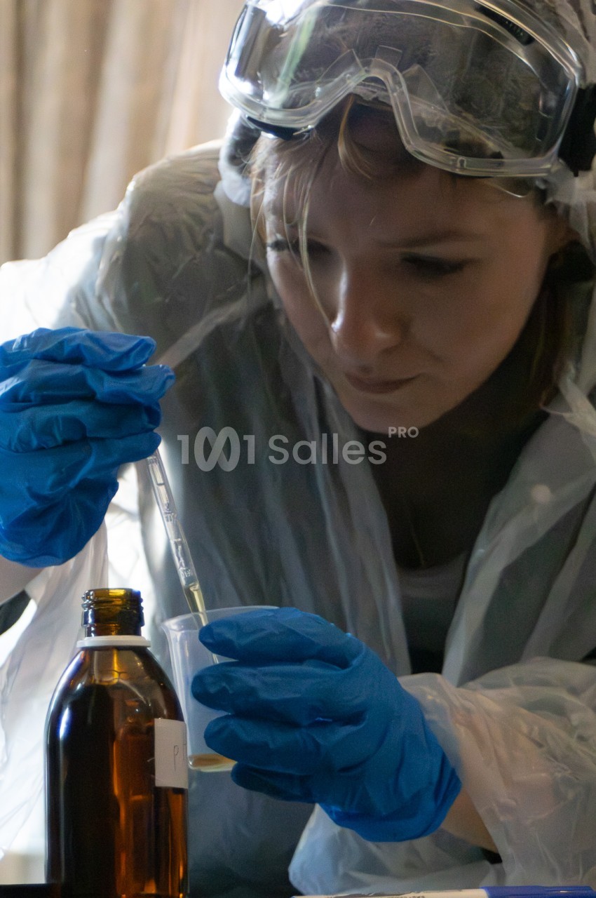 Une personne portant des lunettes de protection, une combinaison blanche et des gants bleus verse un liquide à l'aide d'une pipette dans un bécher, à côté d'un flacon brun.