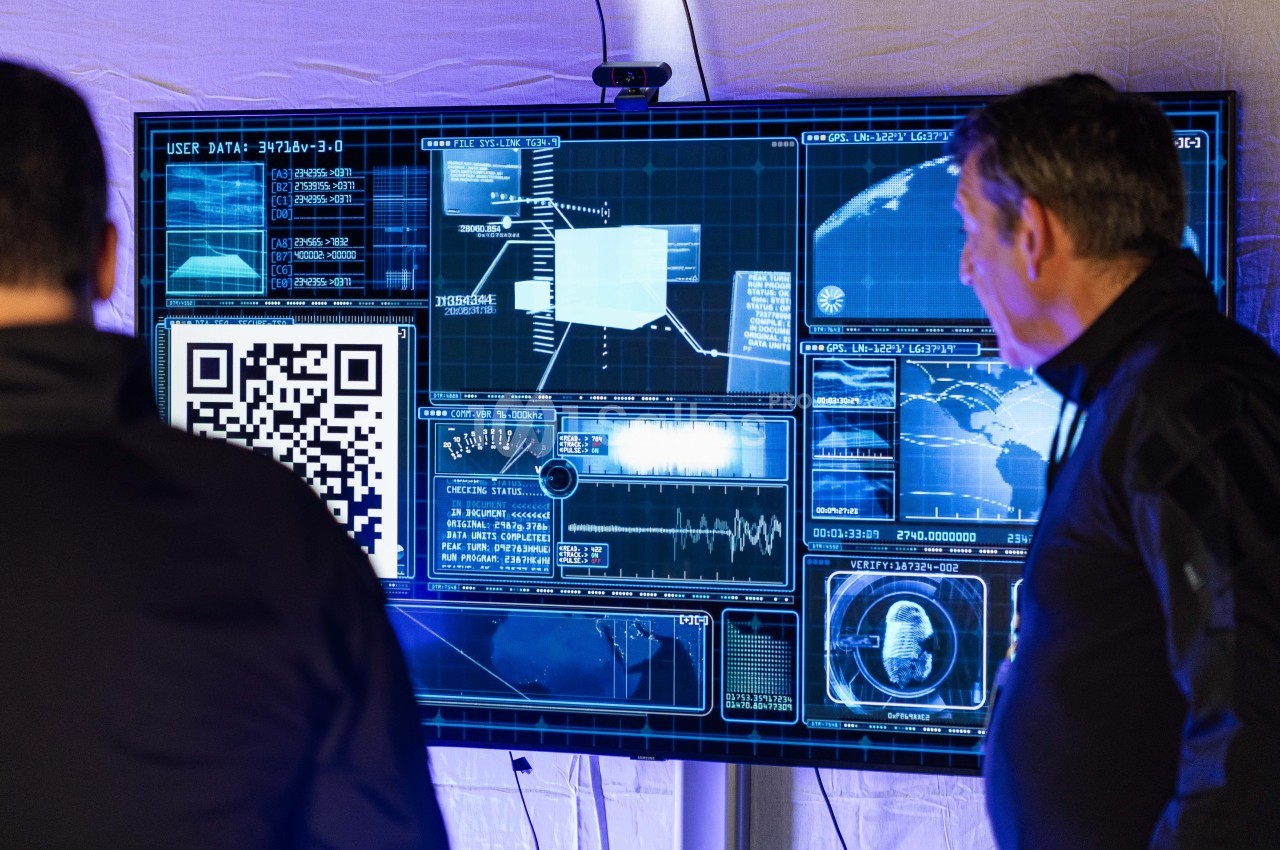 Deux hommes de dos et de profil observent un grand écran affichant une interface futuriste bleue avec cartes, graphiques, code et un QR code au premier plan.
