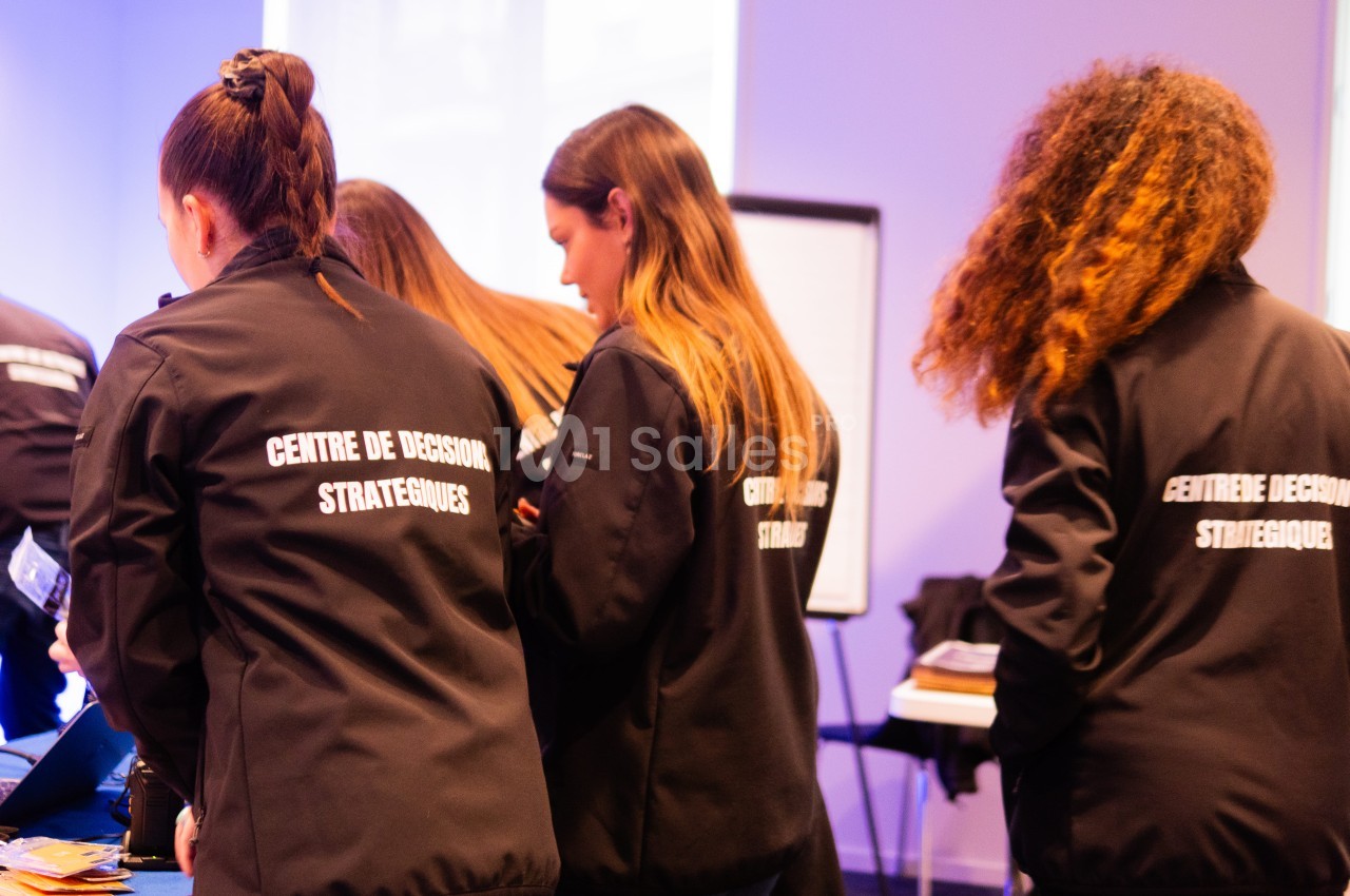 Trois jeunes femmes de dos, vêtues de vestes noires portant l'inscription « Centre de décision stratégiques », observent un plan de travail lors d'un événement en salle. Wait, I must respect the rules: no quotes around? The rule says no guillemets