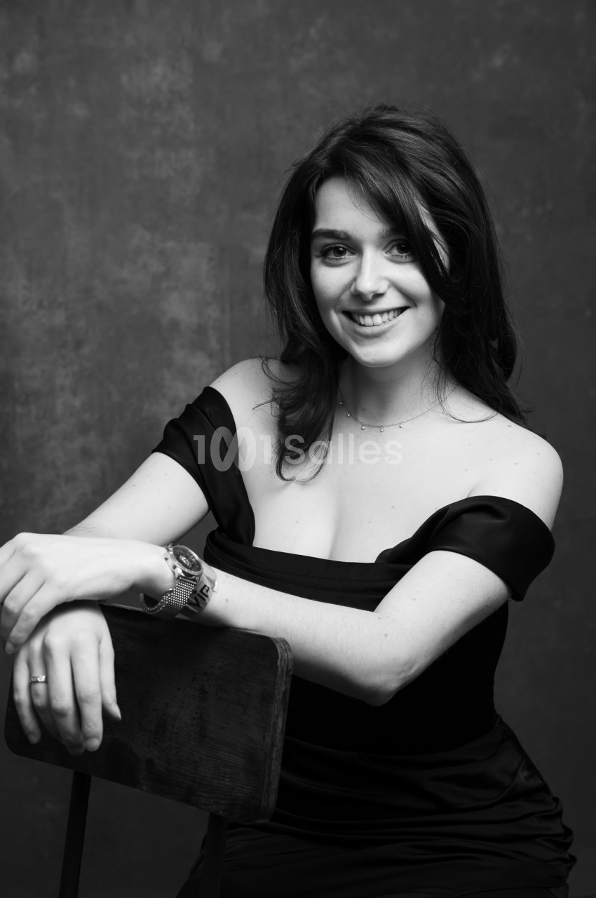 Portrait en noir et blanc d'une femme souriante, assise de profil sur une chaise en bois, bras posés sur le dossier, devant un fond gris texturé.