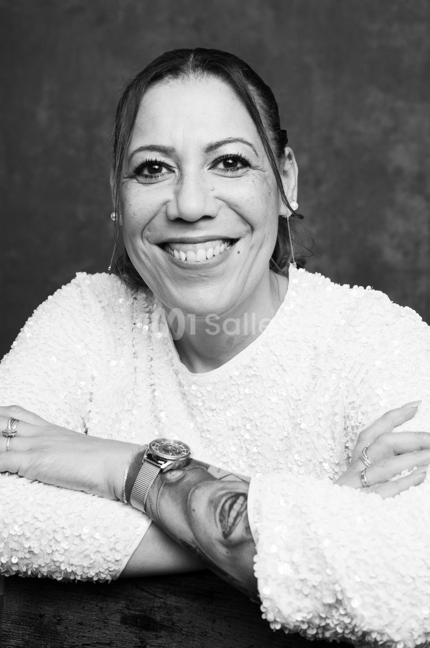 Portrait en noir et blanc d'une femme souriante, bras croisés sur une table, vêtue d'un haut à sequins blancs, sur fond sombre uni.