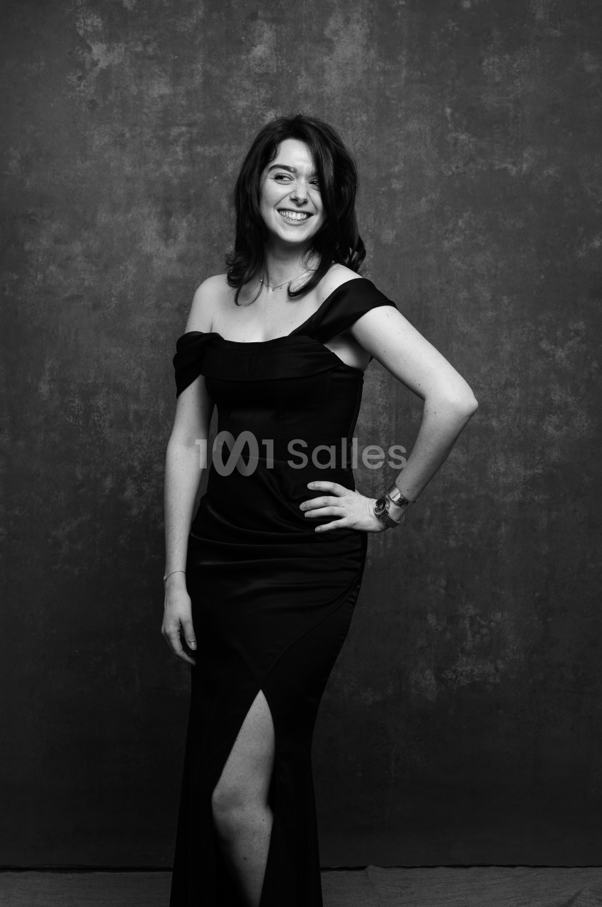 Portrait en noir et blanc d'une femme souriante, debout en robe longue noire à bretelles et fente, une main sur la hanche, devant un fond gris texturé. Hmm, check length: roughly 170 characters, too long. Shorten. Portrait en noir et blanc d'une femme