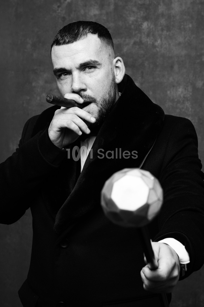 Homme en noir et blanc, manteau sombre à col large, fumant un cigare et tenant un objet sphérique facetté au premier plan, sur fond gris texturé.