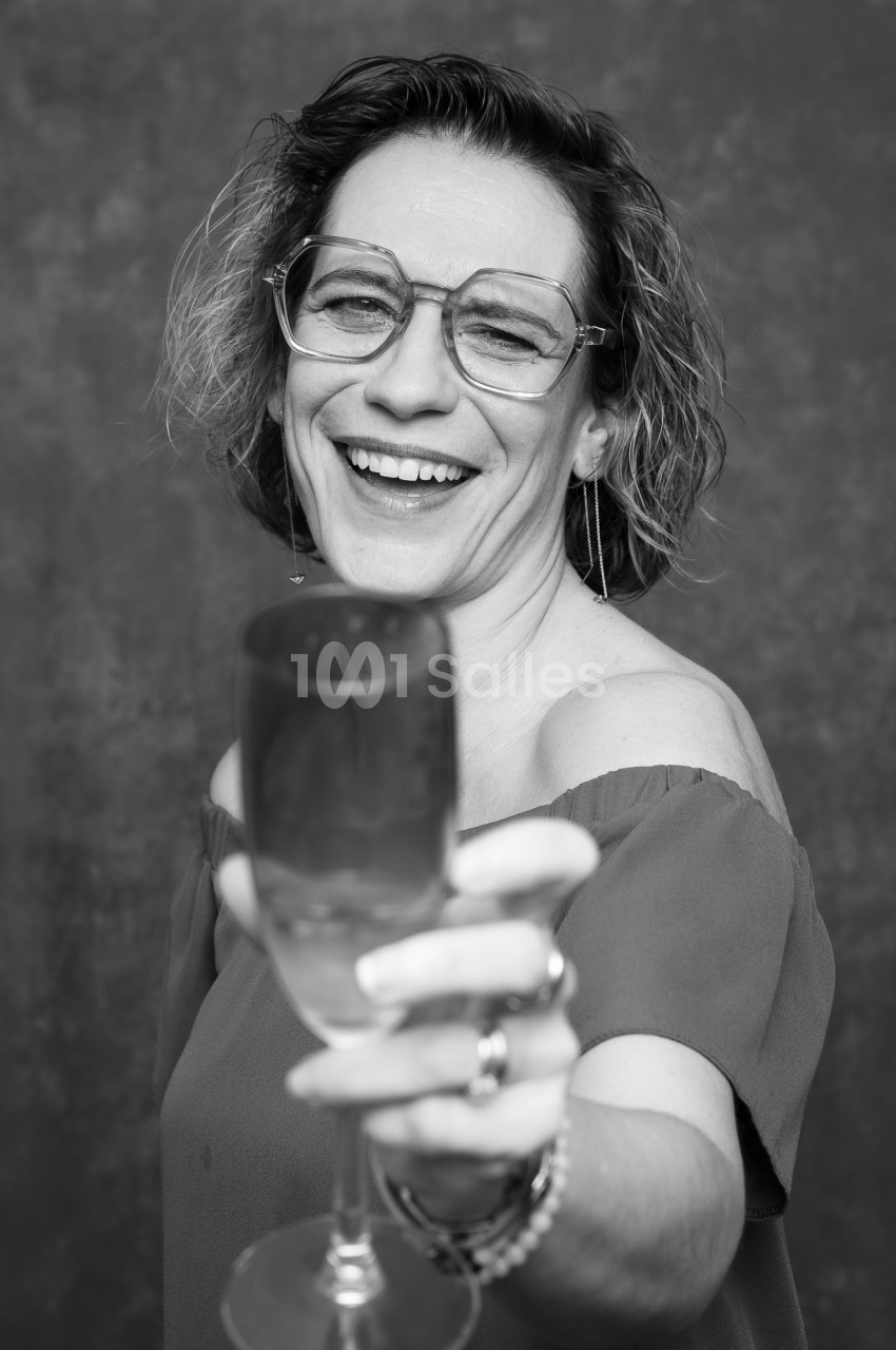 Portrait en noir et blanc d'une femme souriante aux lunettes transparentes, tenant un verre à pied au premier plan vers l'objectif.