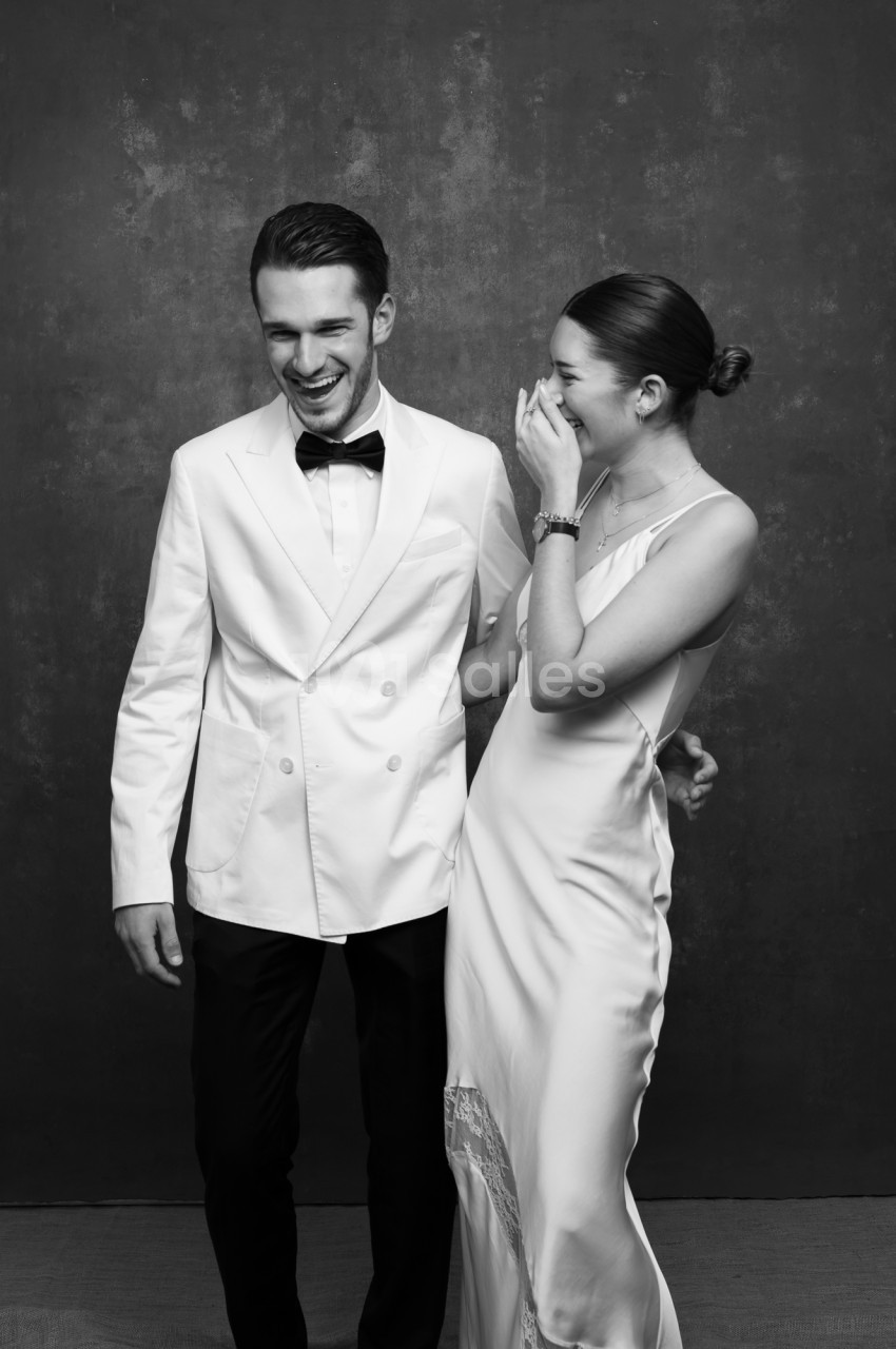 Deux personnes en noir et blanc posent devant un fond sombre texturé : un homme en veste blanche et nœud papillon rit, une femme en robe fluide blanche rit en portant la main à son visage. Let me check length: roughly 180 characters, too long. Need ≤125.