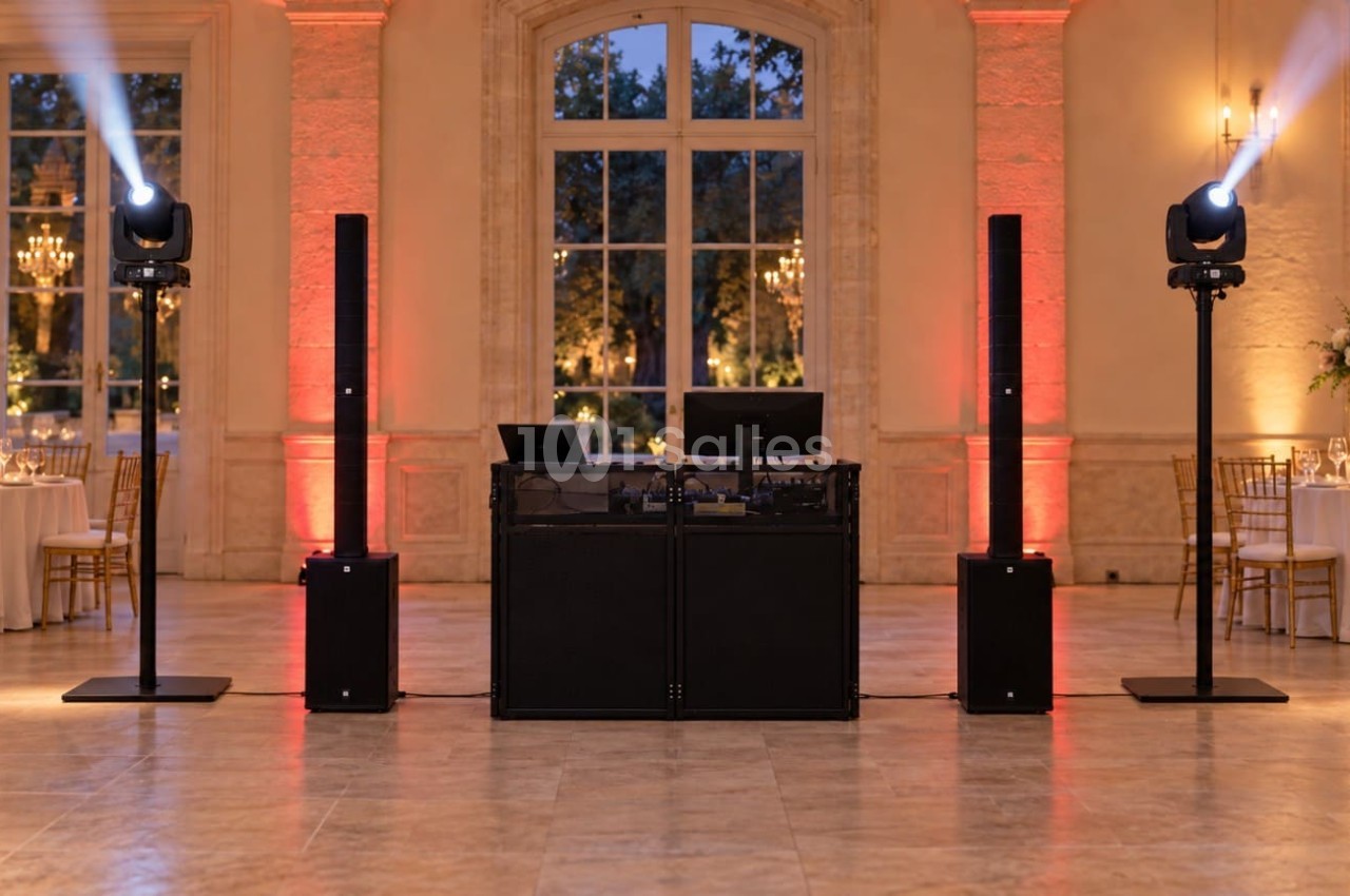 Installation de DJ en salle de réception : console centrale noire encadrée de deux colonnes d'enceintes, projecteurs à tête mobile sur pieds, murs éclairés en orangé. Let me check length: roughly 165 characters, too long. Need ≤125. Shorter
