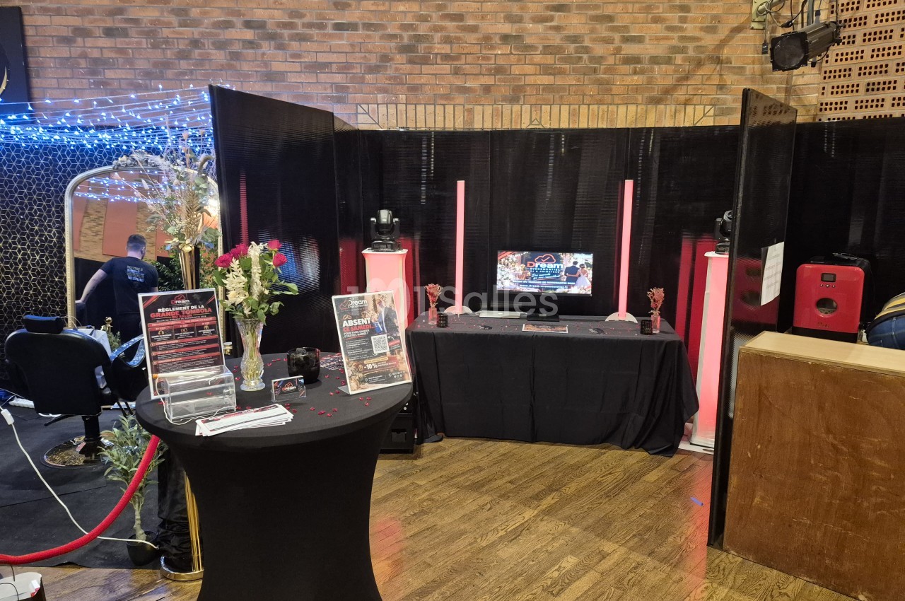 Stand d'animation avec table haute et table basse couvertes de tissu noir, un écran, des bougies roses et un vase de roses, devant des panneaux noirs et un mur de briques. Hmm, need to check length: about 160 characters, too long. Shorten. Stand