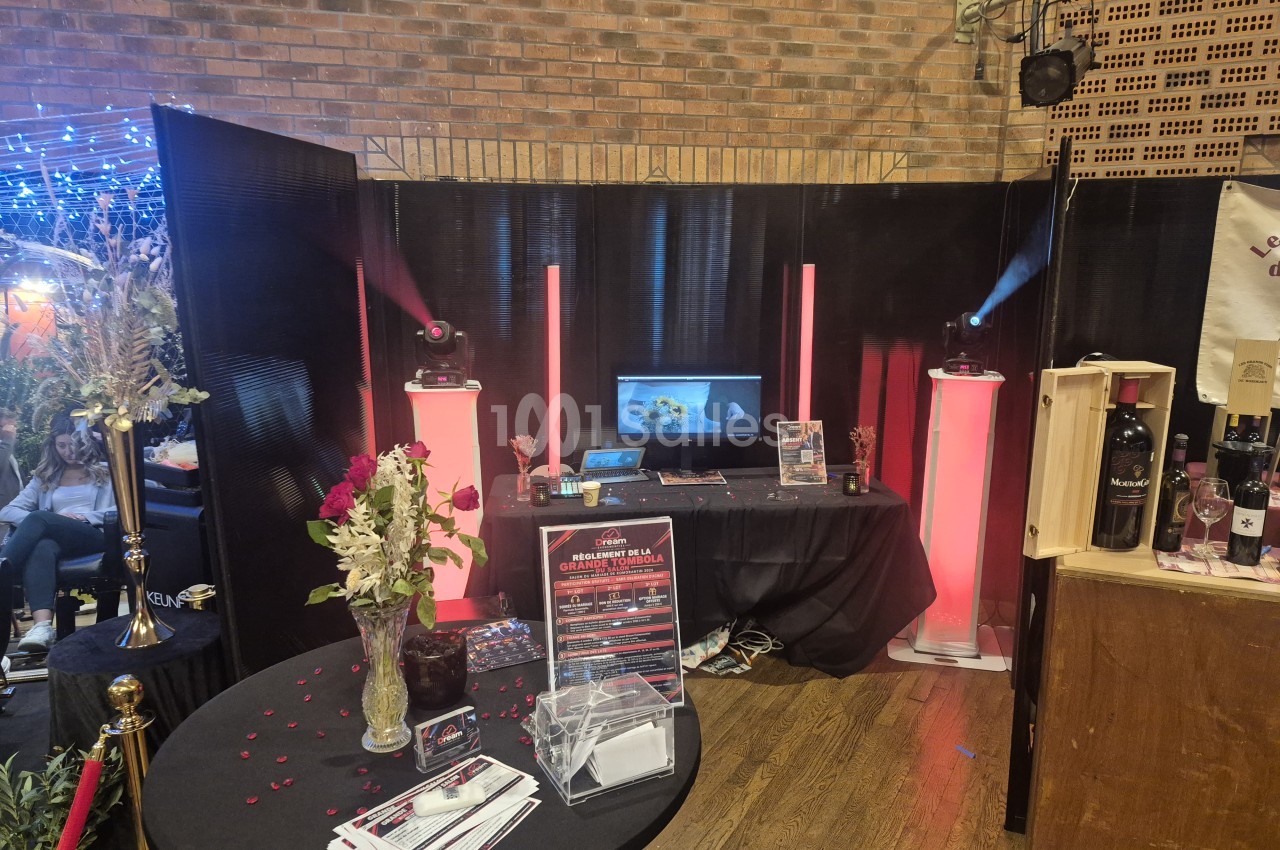 Stand d'animation avec table drapée de noir, ordinateur portable, tubes lumineux roses et deux projecteurs sur pieds, devant un mur de briques et des panneaux noirs.