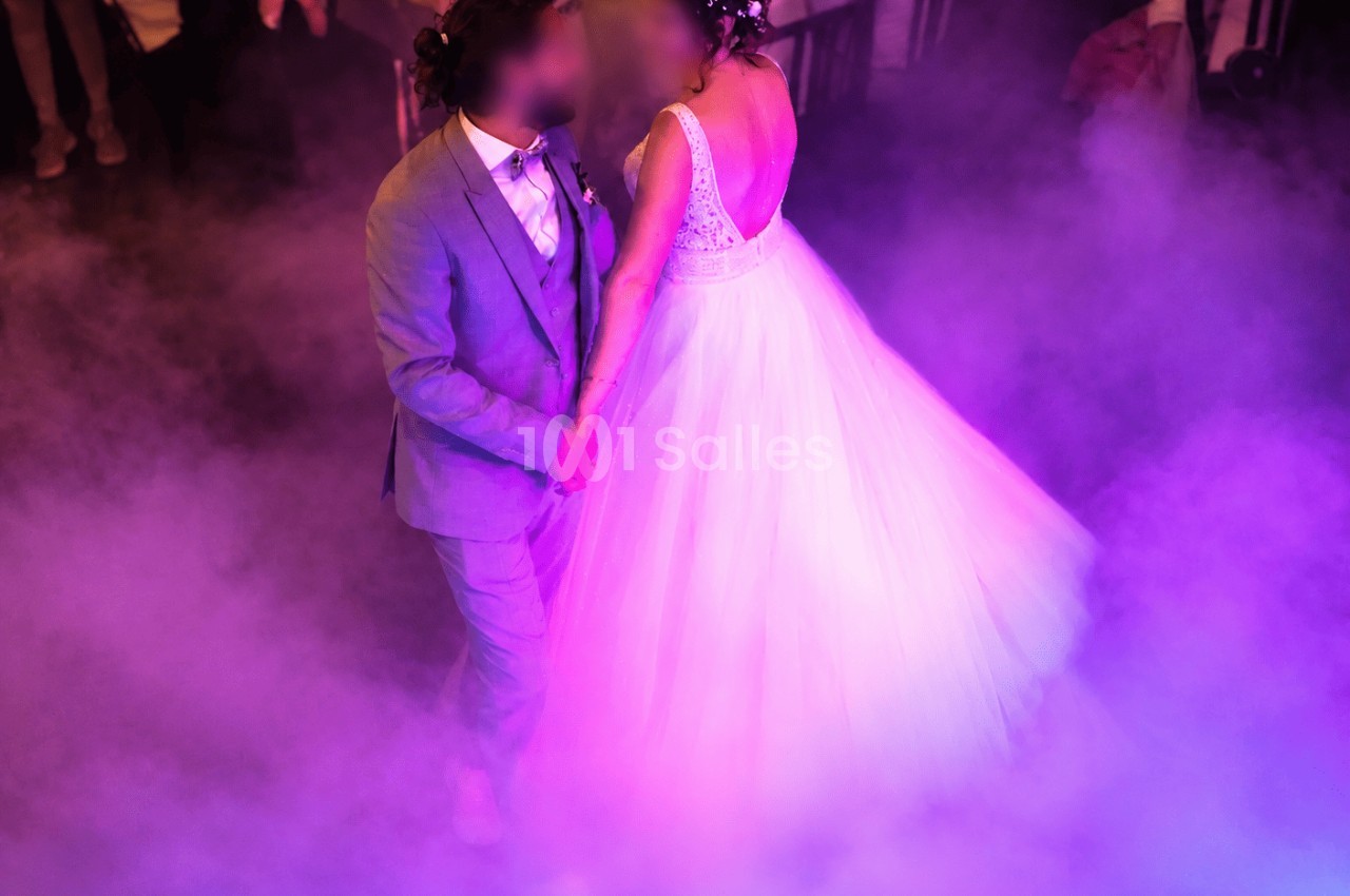 Deux mariés dansent au centre d'une piste enfumée, éclairée en lumières violettes et roses, la mariée en robe blanche à corsage dos nu.