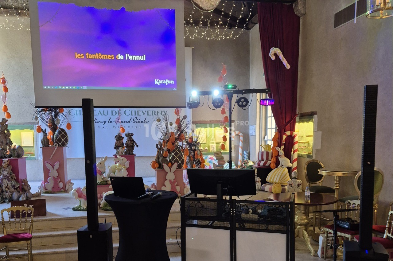 Espace de réception avec un écran de projection affichant des paroles de karaoké, un pupitre DJ et des micros sur une table haute noire, entourés de décors de Pâques.