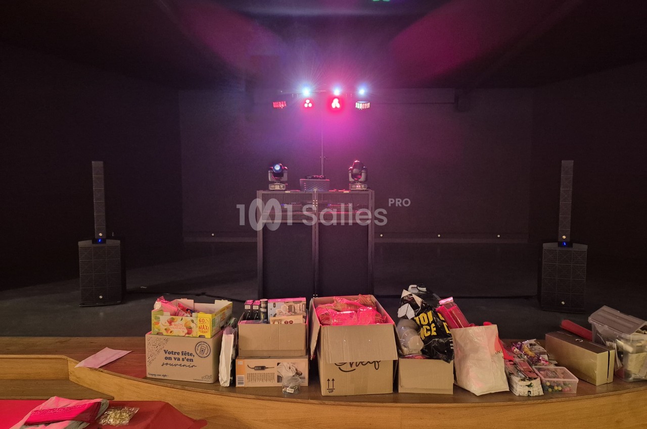 Estrade éclairée en rose et bleu avec table de DJ et enceintes, cartons de matériel de fête au premier plan.
