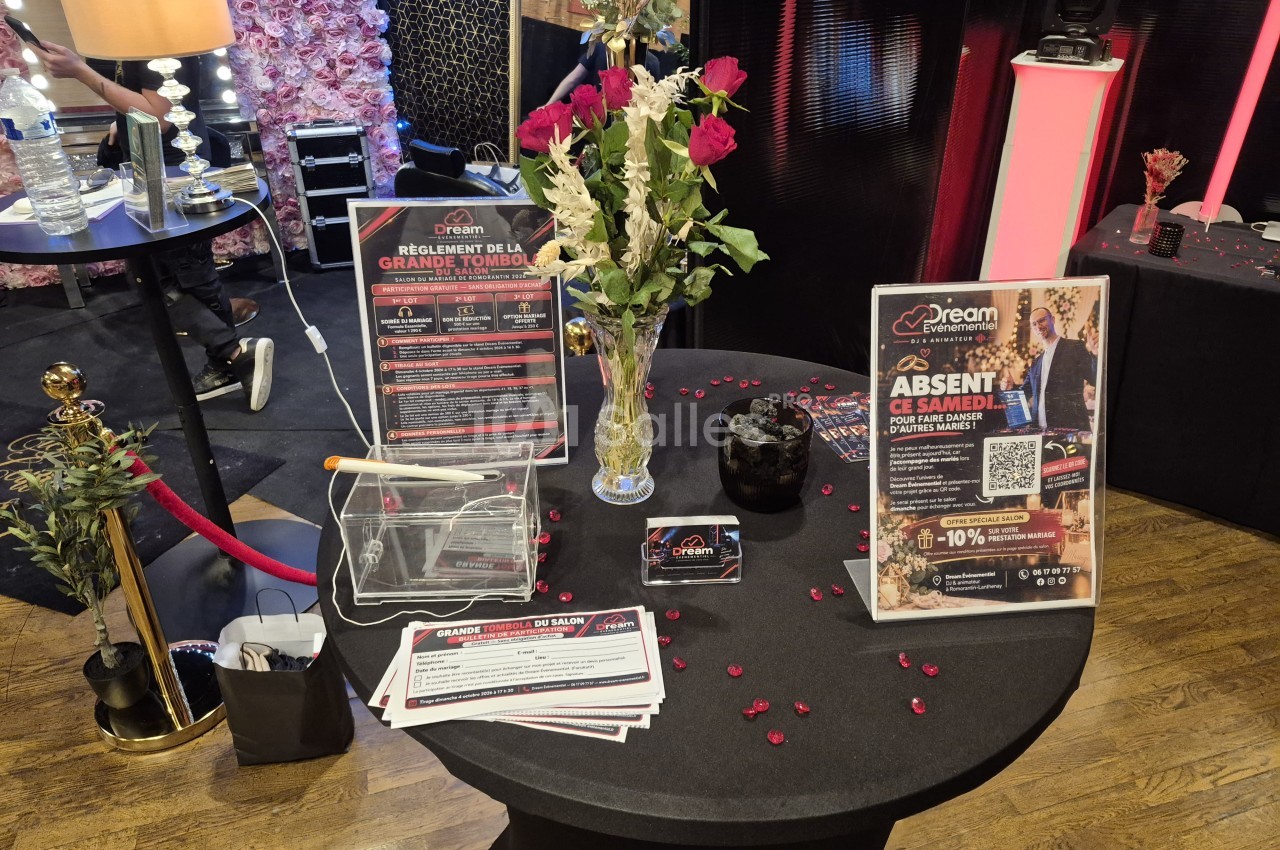 Table ronde noire dressée de roses rouges et blanches dans un vase en cristal, avec des pancartes promotionnelles, une urne transparente et des pétales rouges éparpillés.