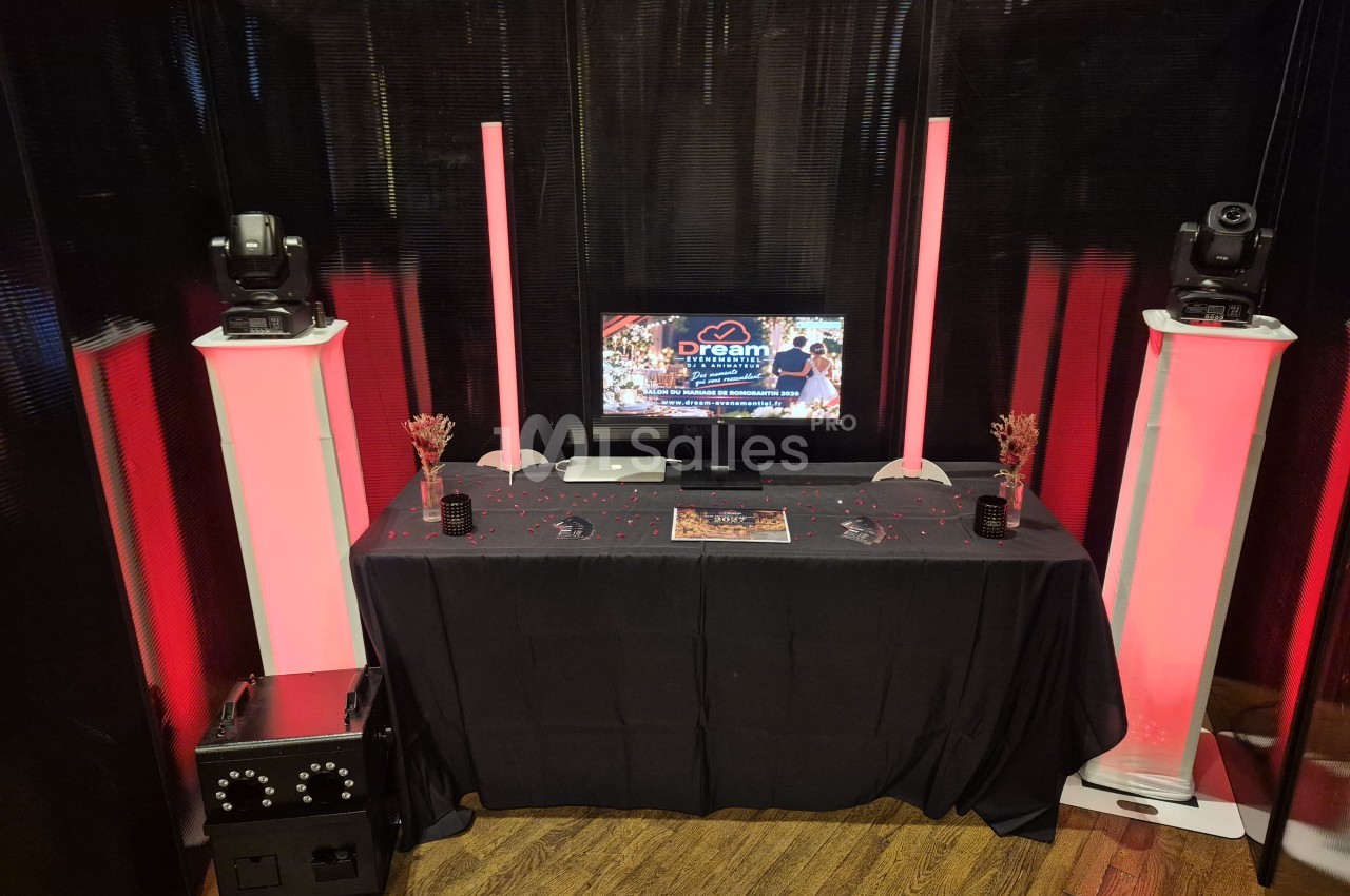 Table noire drapée avec un écran affichant un visuel, flanquée de deux colonnes lumineuses roses et de têtes mobiles sur socles, dans une pièce aux murs sombres.