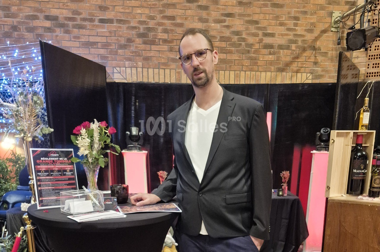 Homme en veste sombre debout près d'une table ronde noire avec un règlement affiché, devant des colonnes roses.