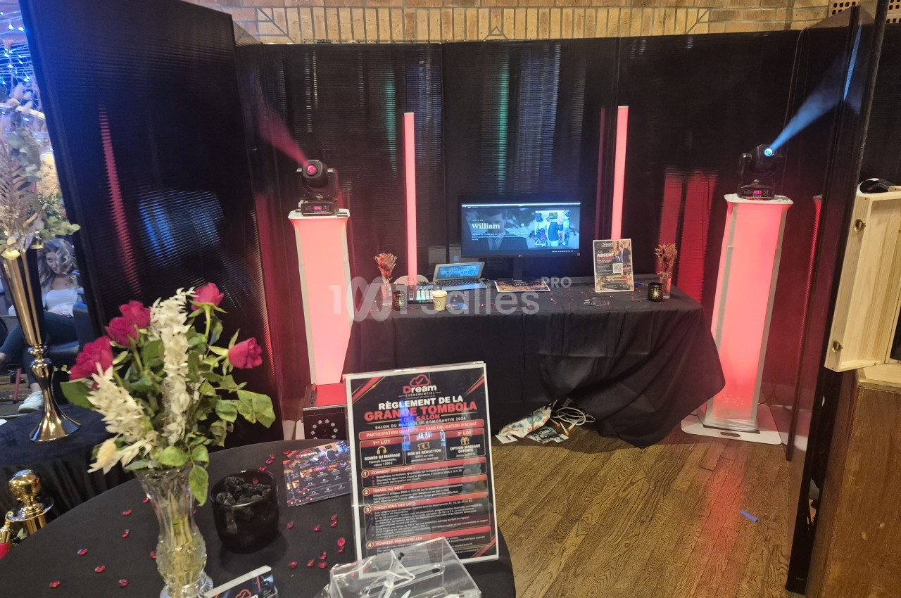 Stand d'animation musicale avec table couverte de tissu noir, ordinateur et écran, deux colonnes lumineuses roses et projecteurs lumineux sur fond sombre, dans une salle aux murs de briques.