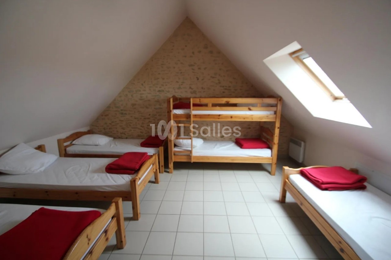 Chambre mansardée avec murs en pierre apparente, deux lits superposés en bois et plusieurs lits simples aux couvertures rouges, sous une fenêtre de toit.