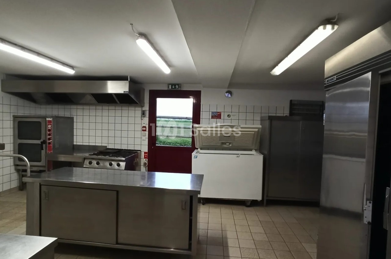Cuisine professionnelle aux carrelages blancs et au sol carrelé, équipée d'une grande table centrale et de plans de travail en inox, avec une porte rouge au fond.