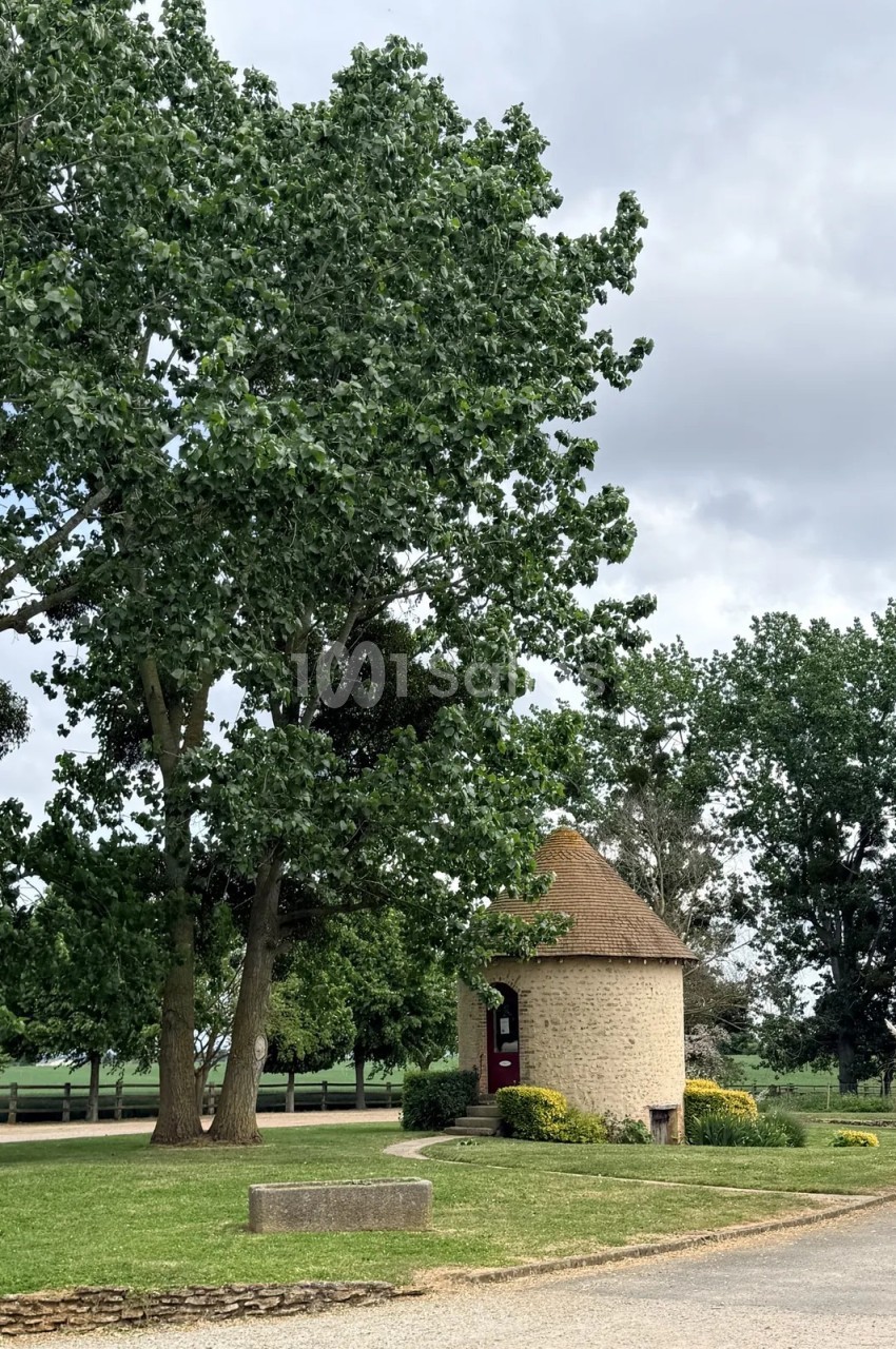 Pigeonnier rond en pierre apparente coiffé d'un toit conique en tuiles, porte rouge, entouré de grands arbres et d'une pelouse, par temps couvert.