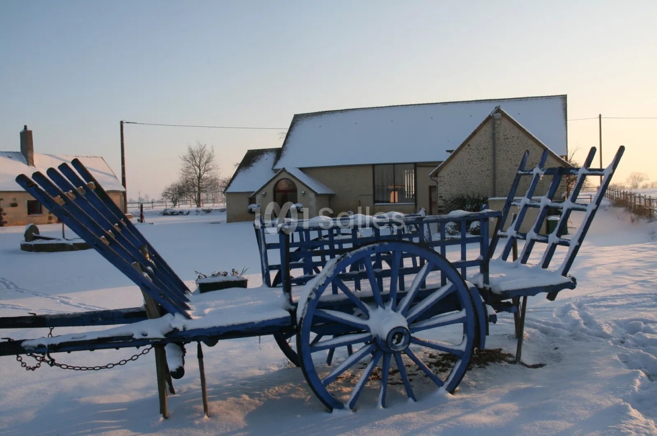 Charrette bleue à roues en bois, couverte de neige, devant un bâtiment de pierre à toit enneigé sous un ciel clair au lever du soleil.