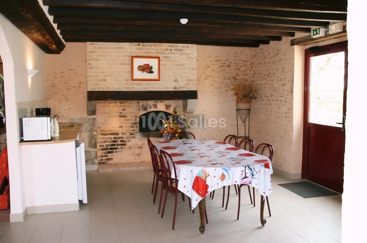 Salle aux murs de pierre et poutres apparentes, avec une grande table recouverte d'une nappe blanche à motifs, des chaises autour et une cheminée en briques.
