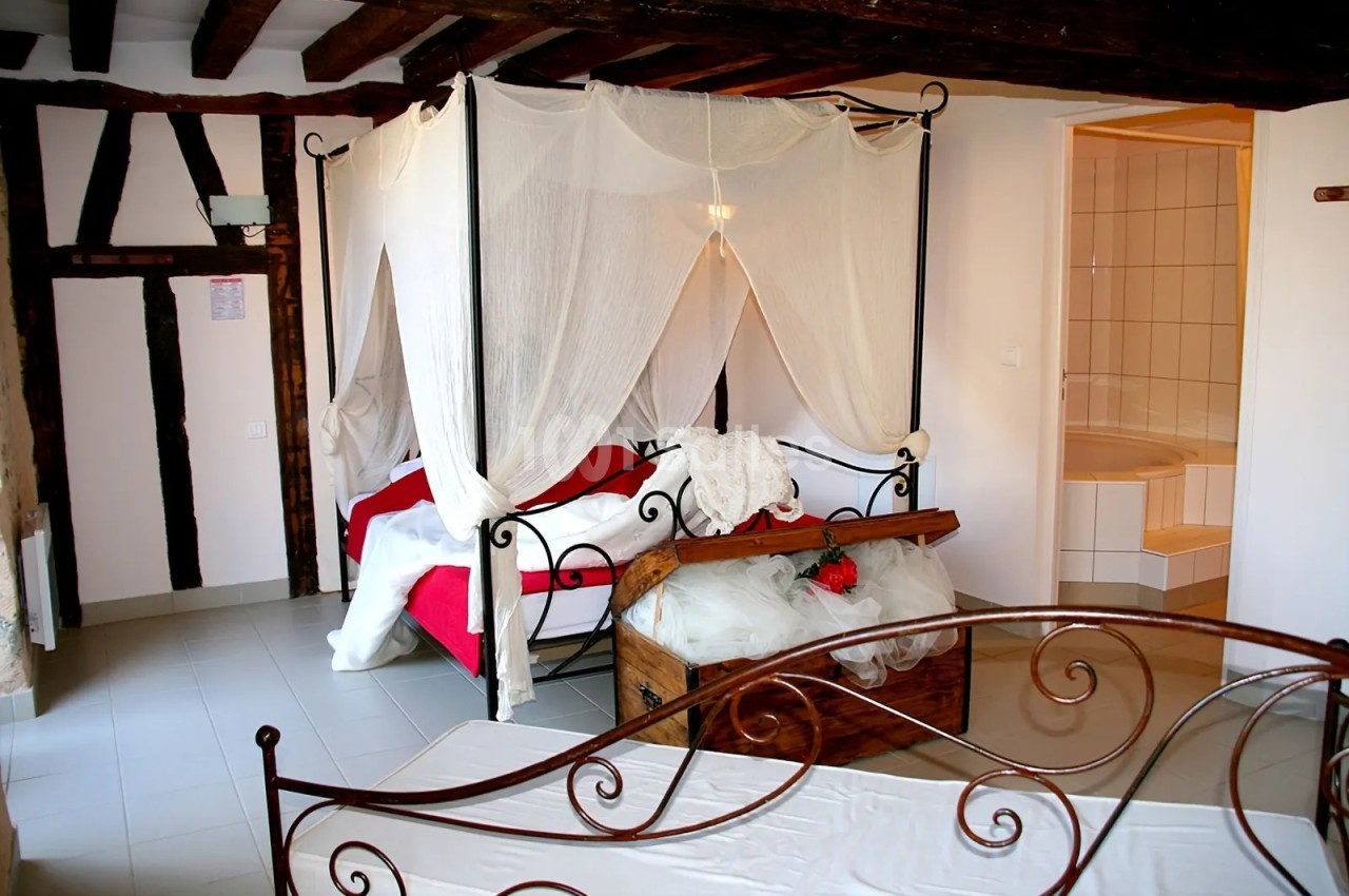 Chambre à thème avec lit à baldaquin en fer forgé couvert de voiles blancs, coffre en bois garni de tulle et de roses rouges, poutres apparentes et colombages.