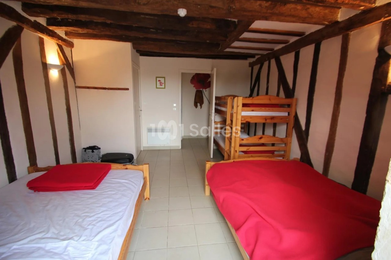 Chambre aux poutres apparentes avec lits en bois recouverts de couvertures rouges, deux lits superposés et un lit simple sur carrelage clair. Let me check length: roughly 135 characters? Count: "Chambre aux poutres apparentes avec lits en bois recouverts